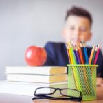 Schoolboy smiling behind a desk with books, pencils, and an alarm clock symbolizing study and creativity.