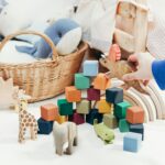 A child's hand arranges colorful wooden toys and animal figures on a playroom floor.