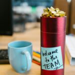 Red gift on desk with sticky note 'Welcome to the TEAM' beside blue mug, ideal for workplace imagery.